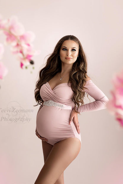 A pregnant woman is wearing a pink bodysuit. The bodysuit is off shoulder and has a sweetheart top. She is wearing a sash right under her breast