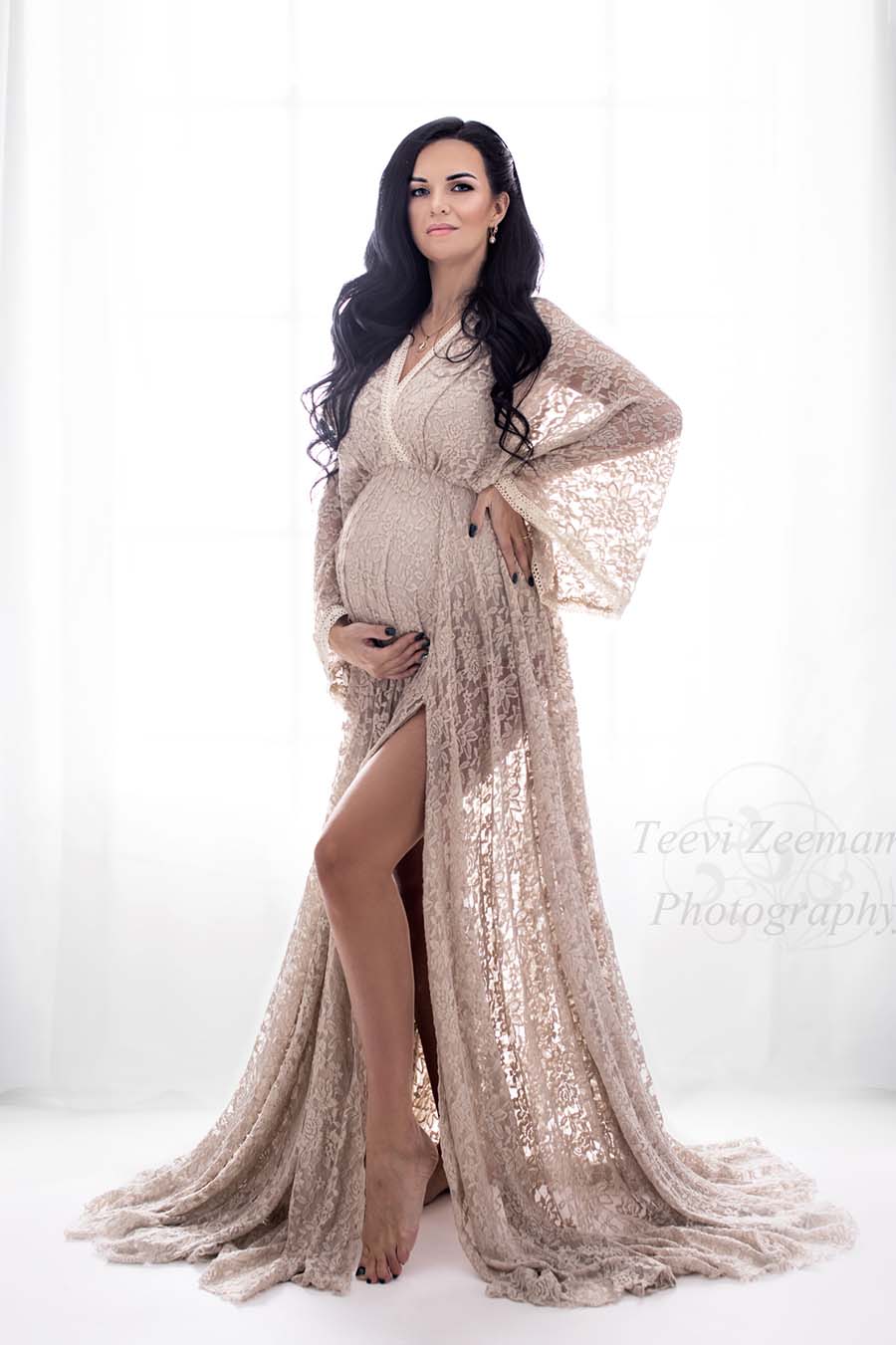 dark haired pregnant model poses in a studio wearing a long dress made of lace in sand color. she stares at the camera while holding her waist and bump. the top of the dress has a low v cut neckline and kaftan sleeves. the long circle skirt has a side split. 