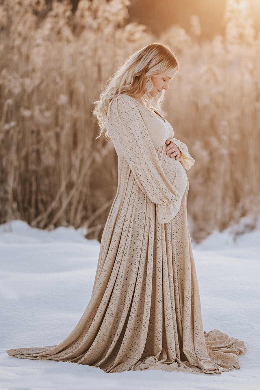 blond pregnant model poses outdoors in the snow wearing a long dress in sand color made of brocante jersey. the dress has poet long sleeves and enough fabric to cover the feet.