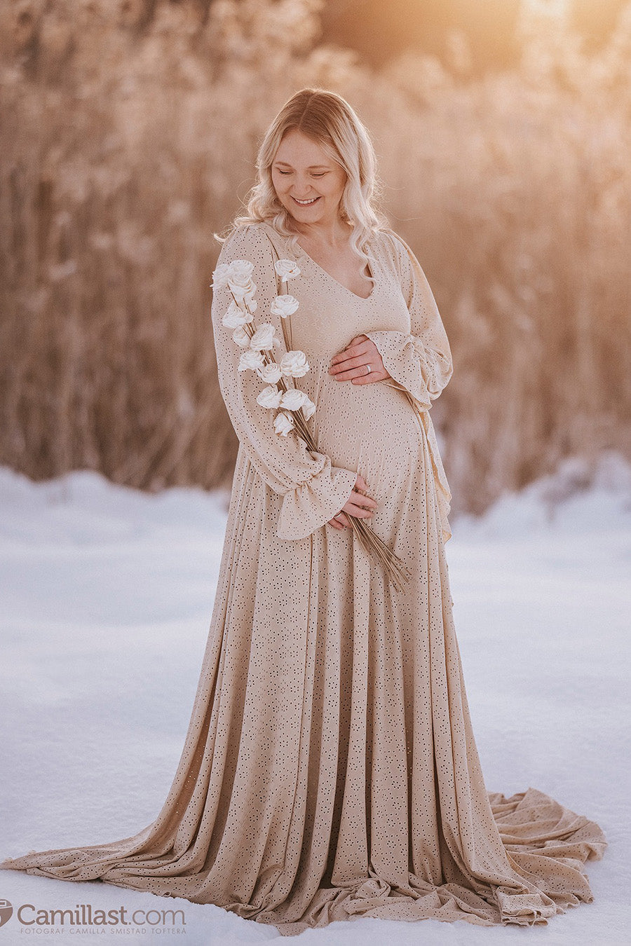 blond pregnant model poses outdoor in the snow wearing a sand long dress made of brocante jersey. the dress features long poet sleeves and a skirt long enough to cover the feet. the model faces down with a smile on her face and holds white flowers.