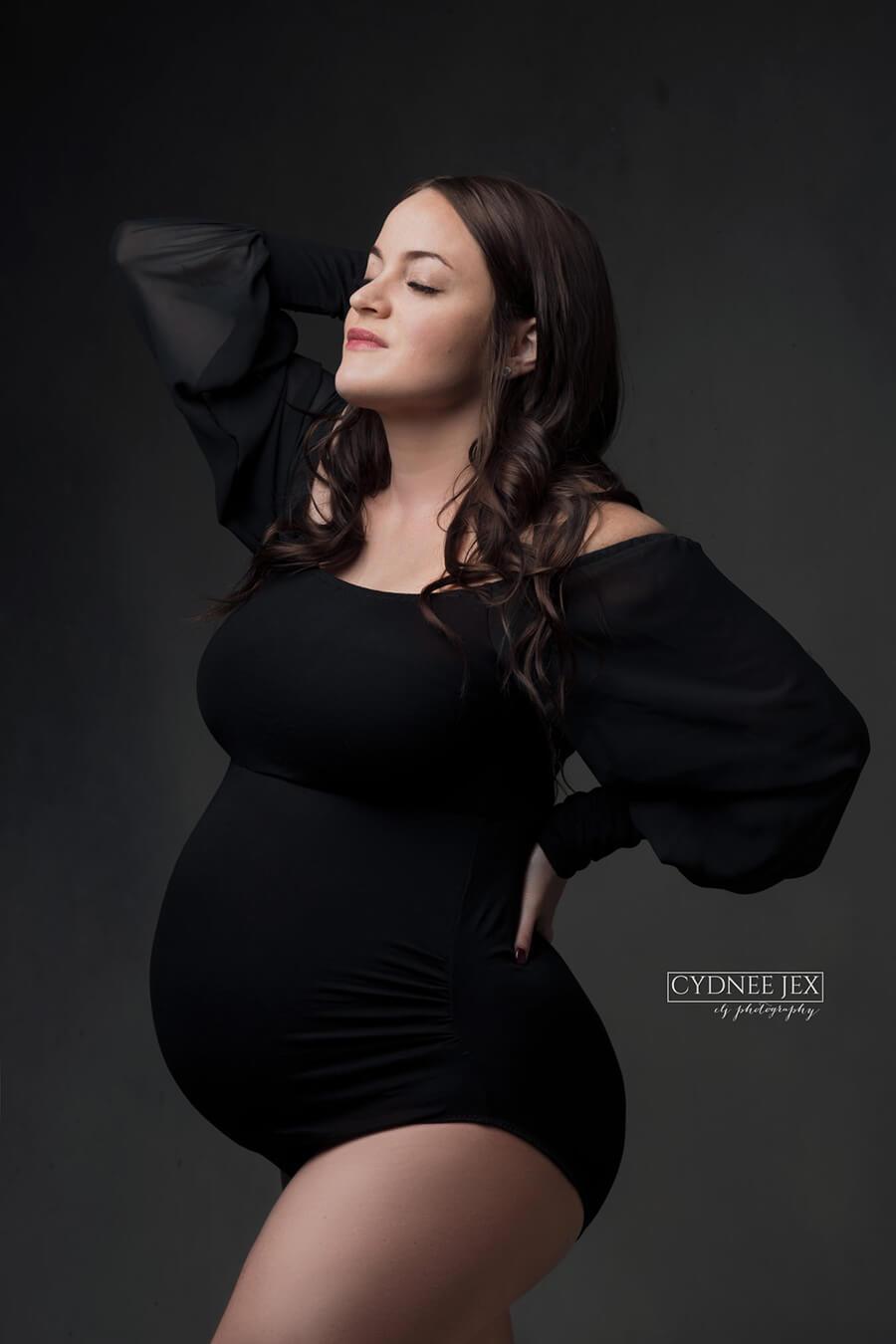 A pregnant woman is wearing a black bodysuit. the bodysuit is off shoulder and has puff sleeves. The sleeves are sheer. She is looking up