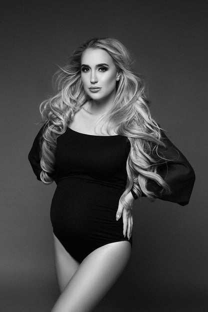A black and white photo of a pregnant woman. She has long curly hair. She is wearing a black off the shoulder bodysuit