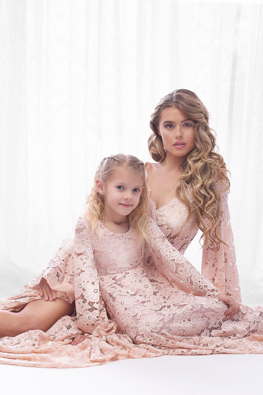 Mother and daughter posing for a mommy and me photoshoot with matching dresses. They are both sitting on the studio floor and wearing a dusty pink dress made of lace. Both dresses are long with long sleeves. Mom and daughter stares at the camera. 
