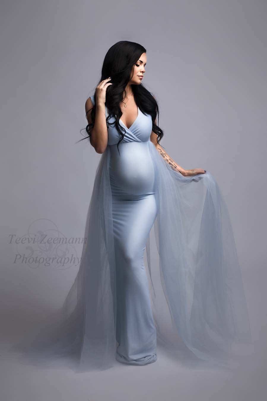 pregnant model wears a light blue dress without sleeves and a adjustable sweetheart top. she has her eyes closed and holds the tulle train from the dress with one hand while the other is resting on her hair.