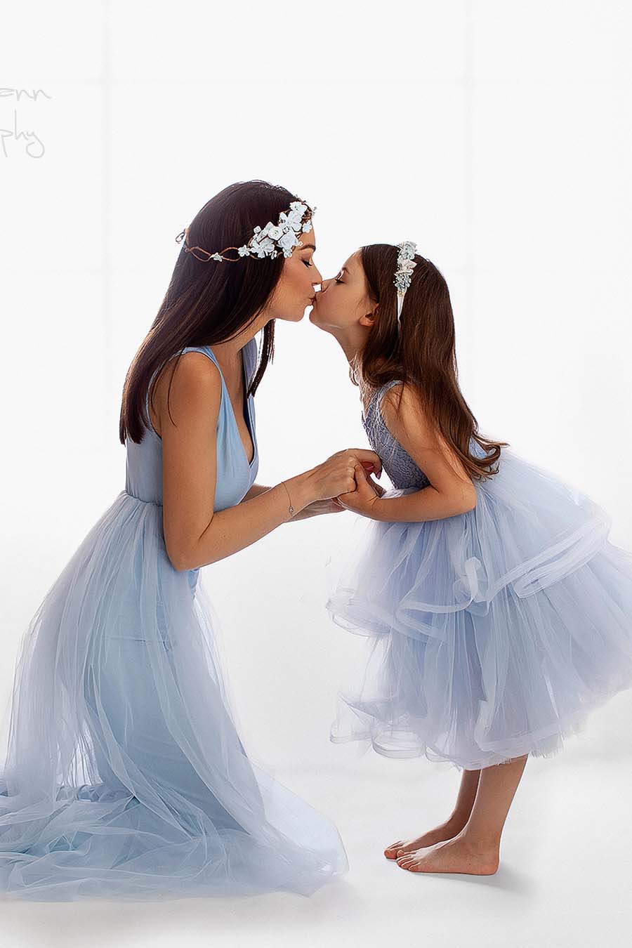 mom and daughter are kissing each other during a close up. they both wear matching outfits without sleeves and tulle skirt. their dresses are light blue.