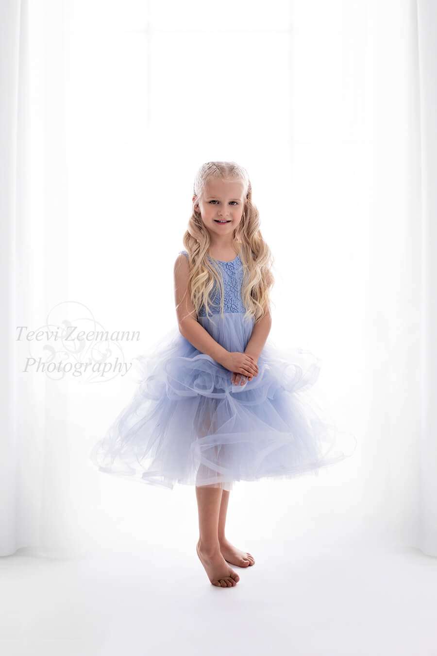 blond kid poses in a white studio wearing a short dress made of lace and tulle in light blue color. she is holding her hands and smiling to the camera.