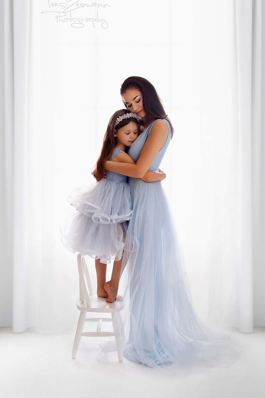 mom and daughter are holding each other during their photoshoot, both wearing matching outfits in light blue. their dresses have no sleeves and a skirt made of tulle.