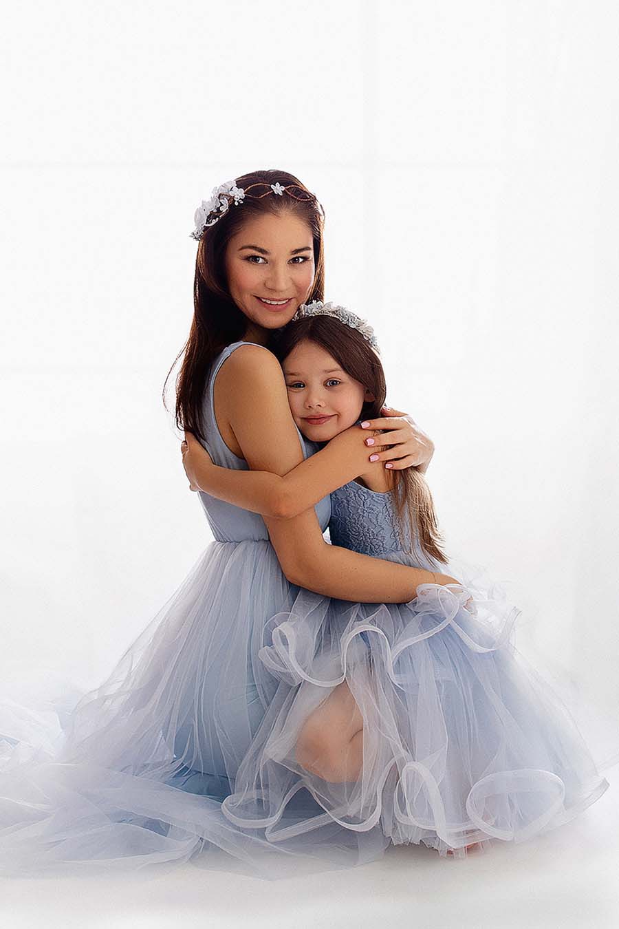 mother and daughter with matching light blue outfits are holding each other during a photo shoot. the dresses have no sleeves and skirts made of tulle. they have both a matching head band.