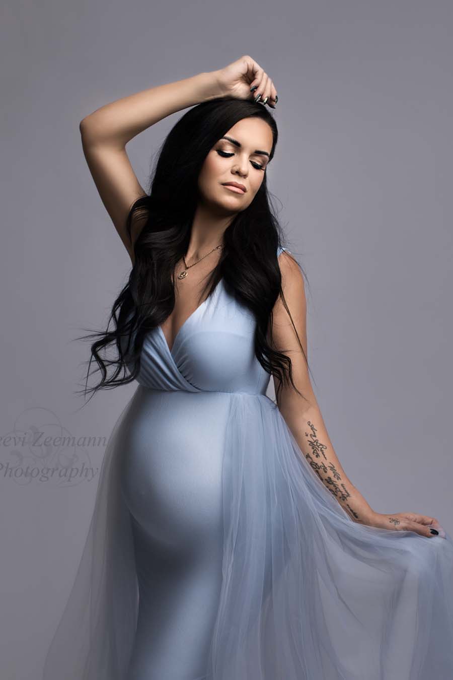 pregnant model wears a light blue dress without sleeves and a adjustable sweetheart top. she has her eyes closed and holds the tulle train from the dress with one hand while the other is resting above her head.