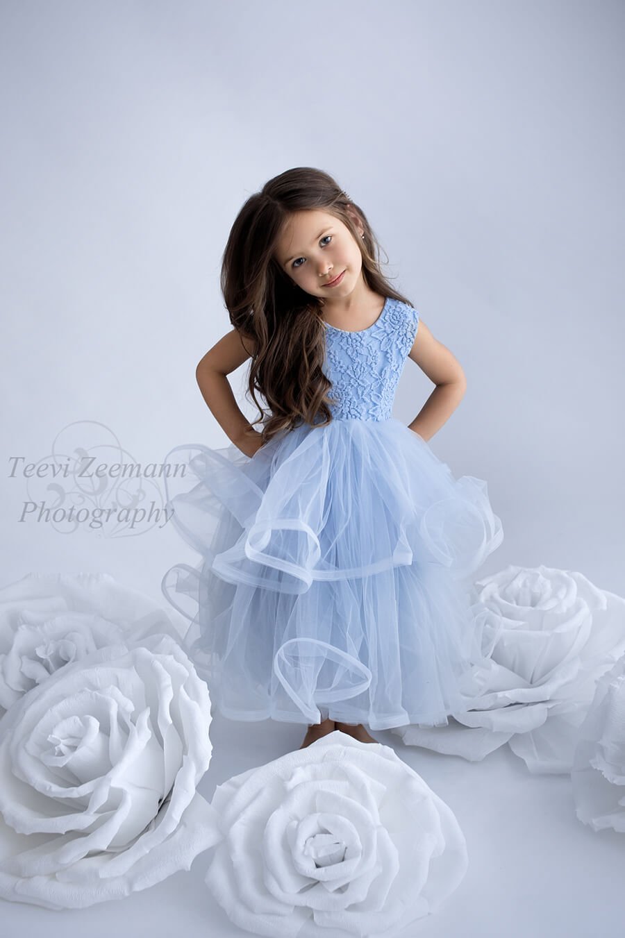 kid wears a light blue dress made of lace and tulle. she has both hands on her waist and the studio is decorated with white big roses.