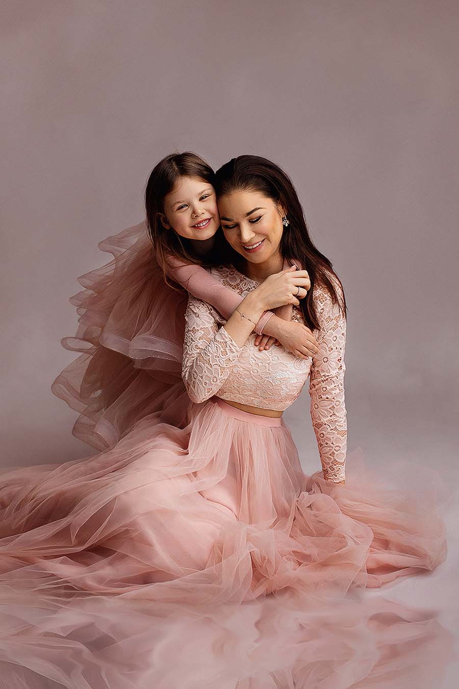 mother and daughter posing sitting on a studio with matching dusty pink outfits with long sleeves and tulle skirt. kid is holding her mom and mom is looking down and smiling. the top of the mother is made of lace while the top from the kid is made of jersey. 