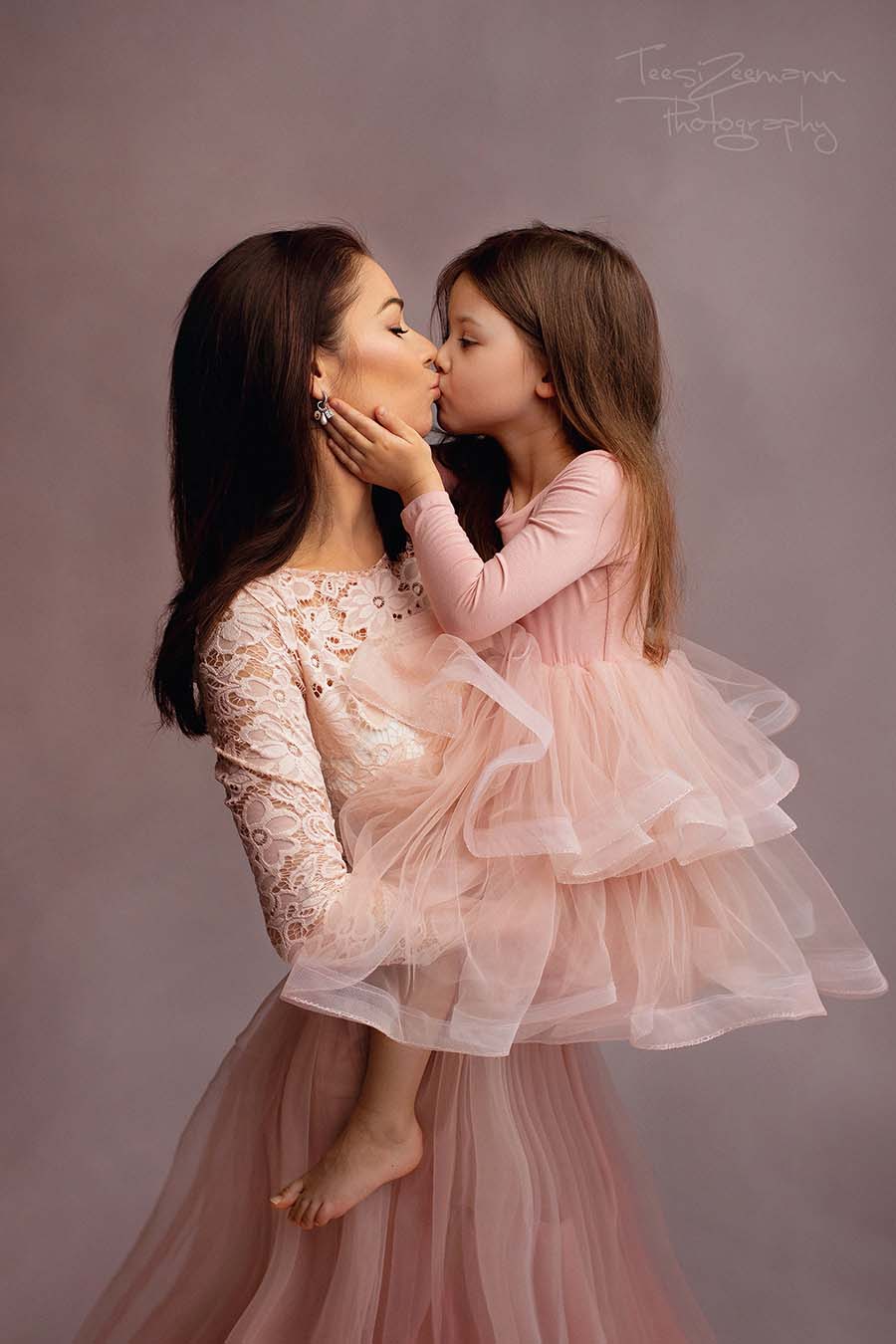 mom and daughter are giving each other a kiss during their photo shoot in a studio. they are wearing matching dresses in dusty pink color. mom has a top with long sleeves made of lace and a tulle skirt while the girl has a top made of jersey and a tulle skirt.