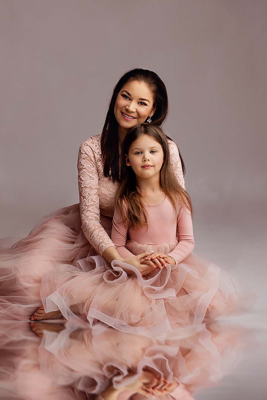 mom and daughter are posing together sitting on the floor during a photo shoot in a studio. they are wearing matching long sleeves dresses in dusty pink color. mom has a lace top and kid has a jersey top. they both have tulle skirt. 