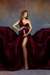 A model with blonde hair is posing in a studio. She is posing in front of a grey background with some orange tints. She is posing with one hand in her side and the other one underneath her babybelly. She is wearing a silky scarf bordeaux as a dress. the dress is one shoulder and has a sash around the waist. She is wearing a matching red headband to finish the look.