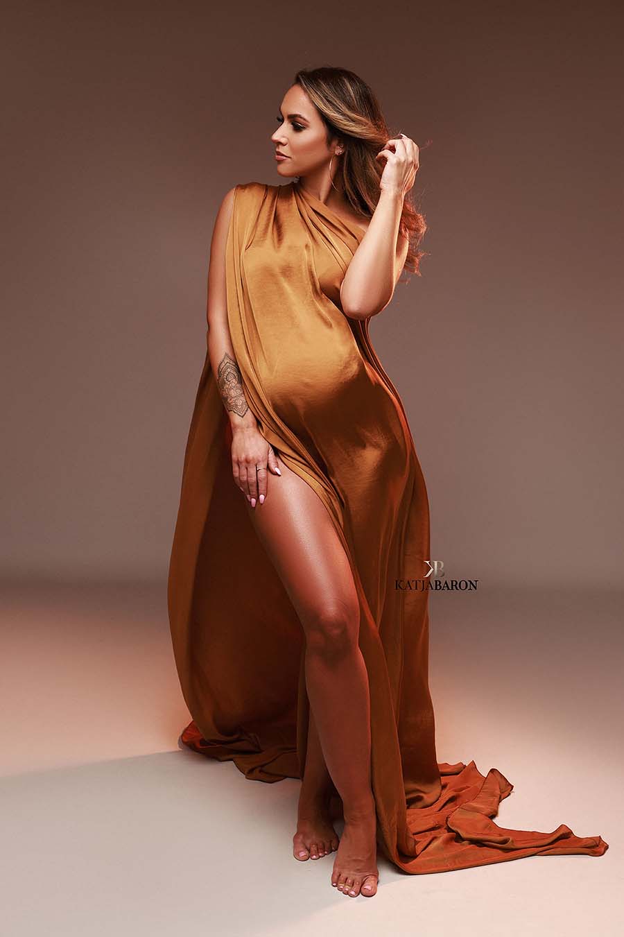 Pregnant model poses in a studio wearing a scarf to cover her body partially. The scarf is cognac camel color and made of silk. She looks to the side and touches her hair.