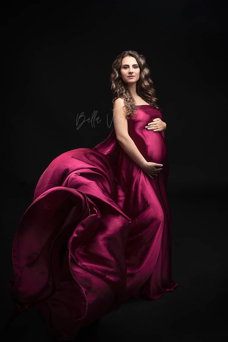 A pregnant woman is wearing a silky scarf cherry around her. She has both her hands around her belly. The fabric at the side of her is tossed in the air. 