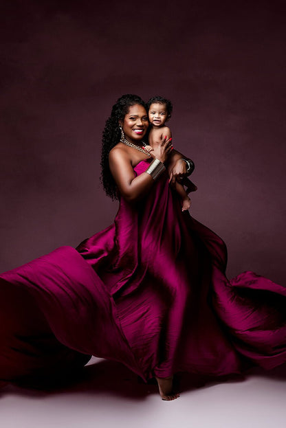 A mother is holding her baby boy. The woman is wearing a dress that is made out of scarf fabric. The color is cherry and the fabric is silky for a shimmer effect. The dress is being tossed around her for a big effect. 