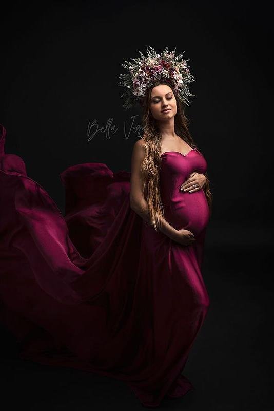A pregnant woman is in a photography studio. She is wearing a silky scarf cherry as a dress. She is posing with her hands around her belly and has her eyes closed. The fabric behind her is being trowed in the air and is slowly falling down. The woman has a big headpiece with lots of flowers on in in her hair.
