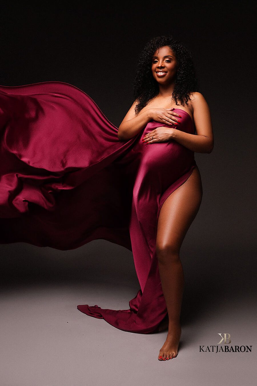 A pregnant model is posing for the camera. They have wrapped the silky scarf around her. Her leg is sticking out of the fabric. The fabric is being tossed in the air.