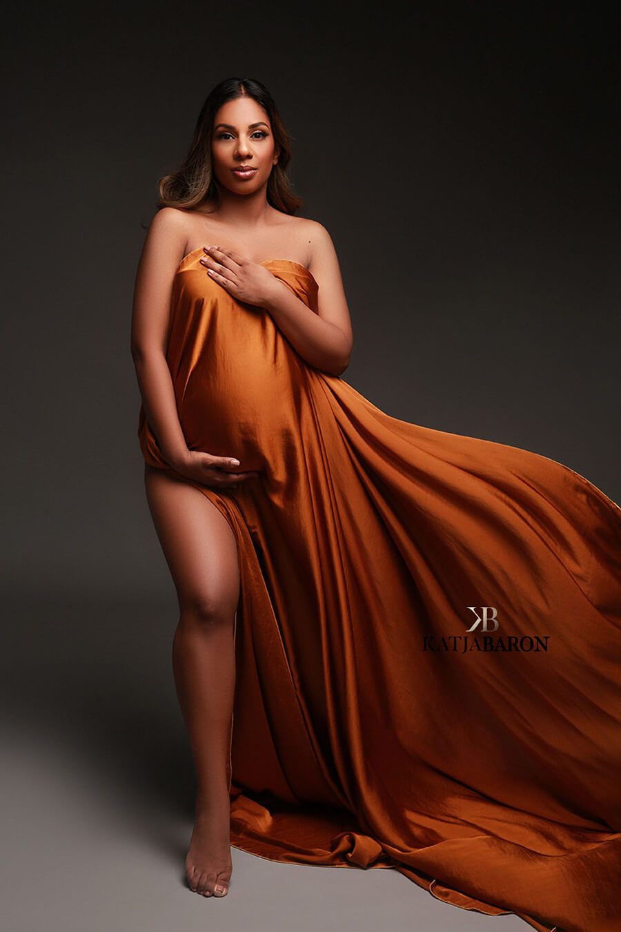 Pregnant model poses in a studio wearing a draping fabric in cognac color. the scarf covers her breast and belly.