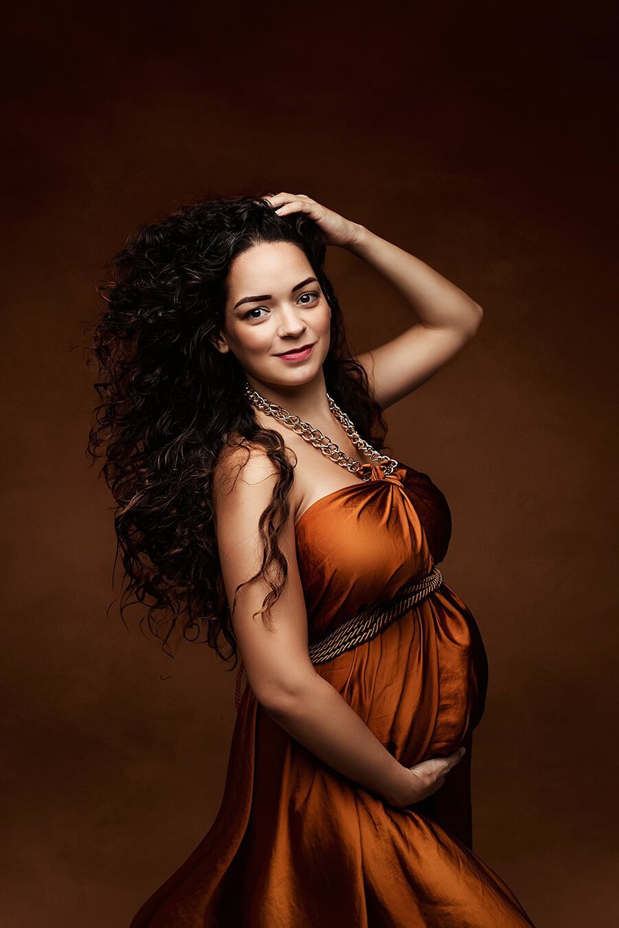 Pregnant model has curly hair and poses on her side with a scarf tied in a necklace. the whole composition looks like a dress. the scarf is made of silk and has a cognac color. 
