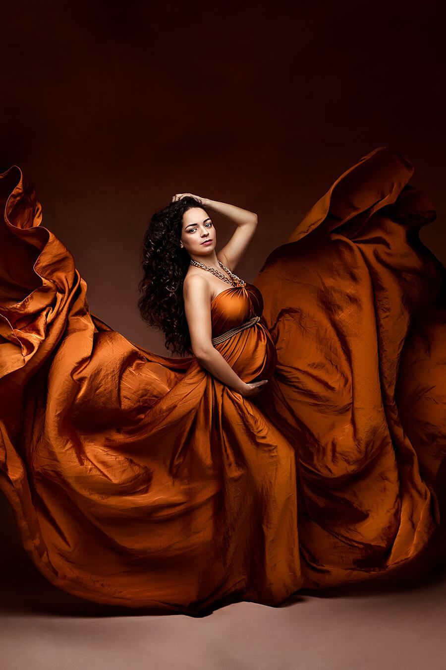 brunette curly model has one hand above her head and the other under her bump. she stares at the camera and wears a cognac draping silk fabric as a dress accessorized with a necklace and a belt.