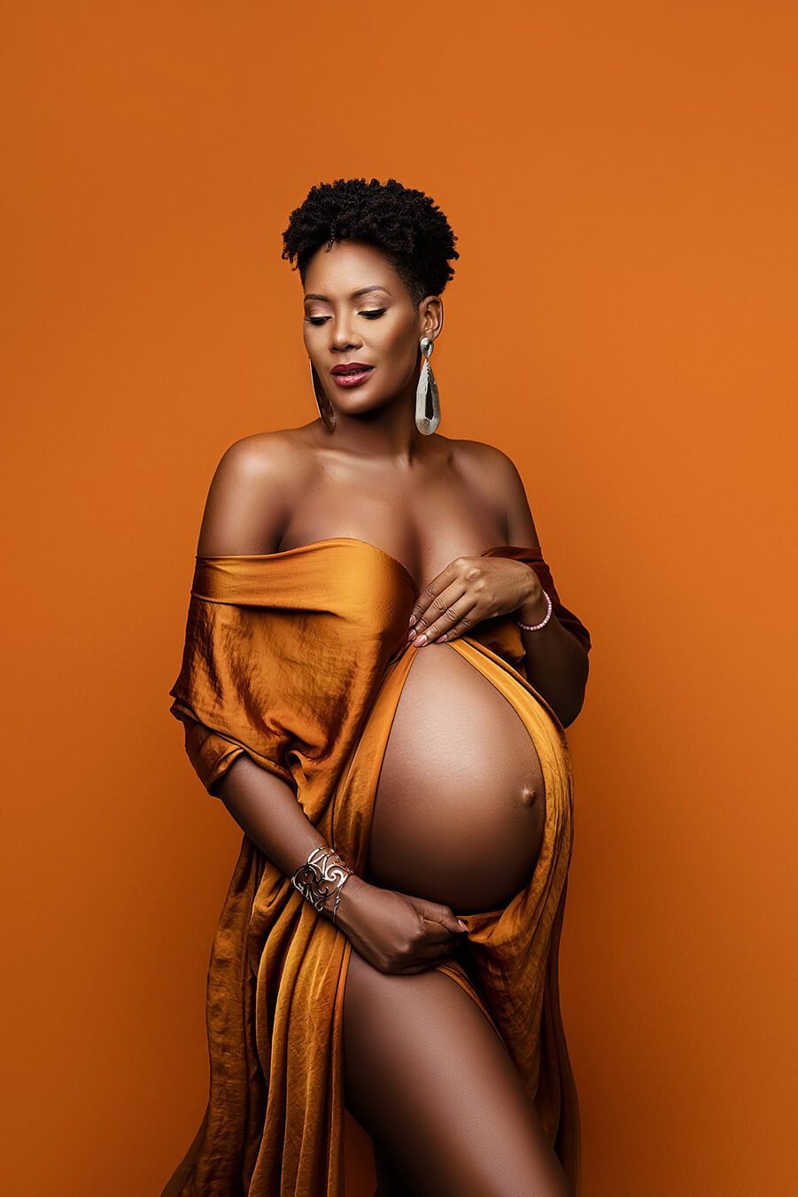 pregnant model poses with a cognac silky scarf wrapped around her arms. she is looking down and poses in front of an orange background with a cognac scarf.