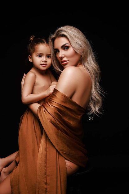 Blond model poses together with her daughter. She has a cognac camel silky fabric wrapped around her arms and they are both staring at the camera.