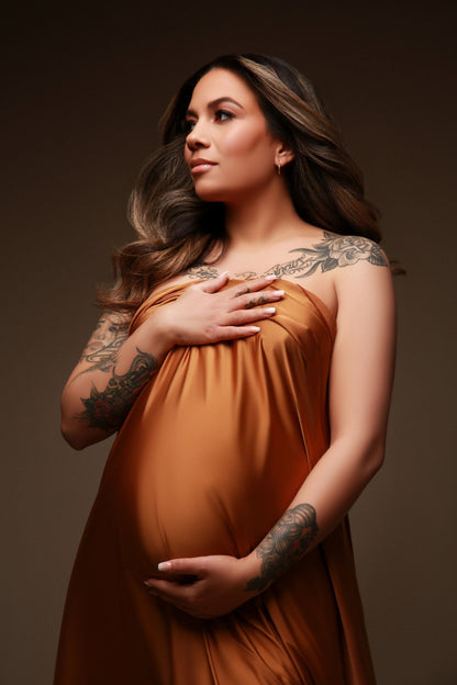 Model is pregnant and covering her body with a cognac camel scarf. She looks to the side and holds the scarf against her belly and chest.
