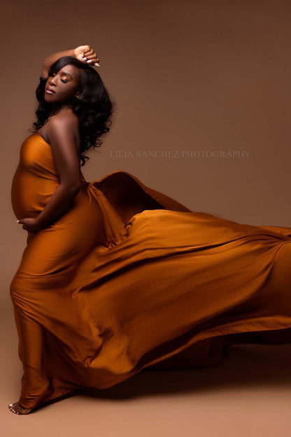 pregnant model poses with her eyes closed in a studio wearing a cognac warm silky scarf to cover her body. she has one arm above her head and the other holds her baby bump.