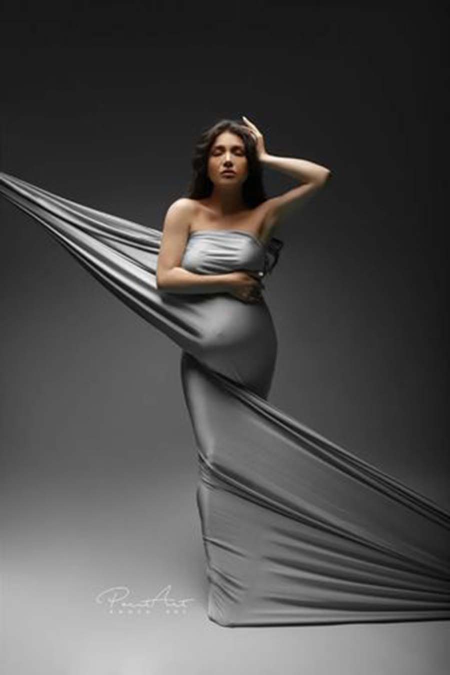 A pregnant woman with long brown hair is posing for the camera. She is being wrapped in a silky scarf in the color cool grey. She has her eyes closed. The two end pieces of the fabric are pulled tight till the end of the photo.