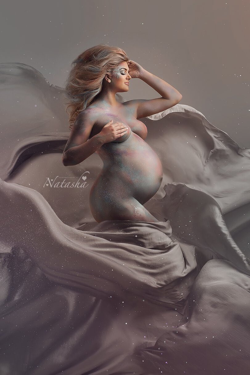 A pregnant model is covered in pastel colored paint. Around her is a silky scarf waving. The scarf is in the color cool grey. It looks like she is floating between the fabric.