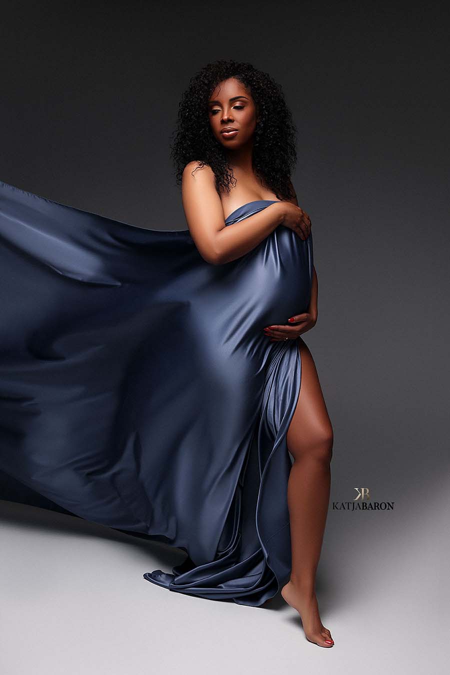 pregnant model with curly hair poses in a studio wearing a piece of draping fabric in denim color covering her chest and belly. one of her legs can be seen. she is looking to the side.