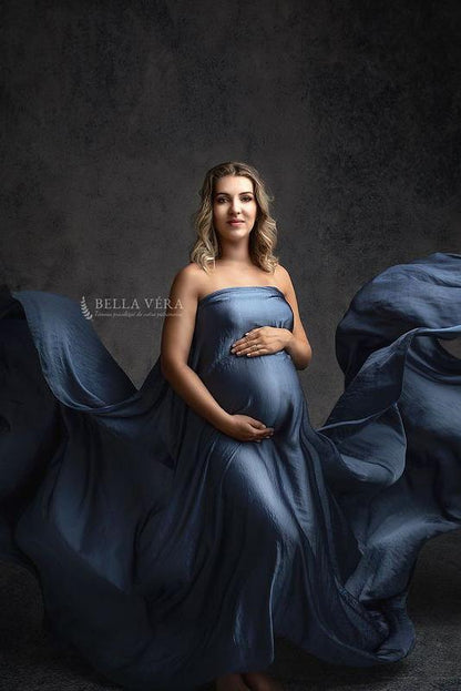 blond pregnant model poses in a studio wearing a piece of draping fabric covering her chest, belly and legs. the fabric is made of silk and the model is staring at the camera.