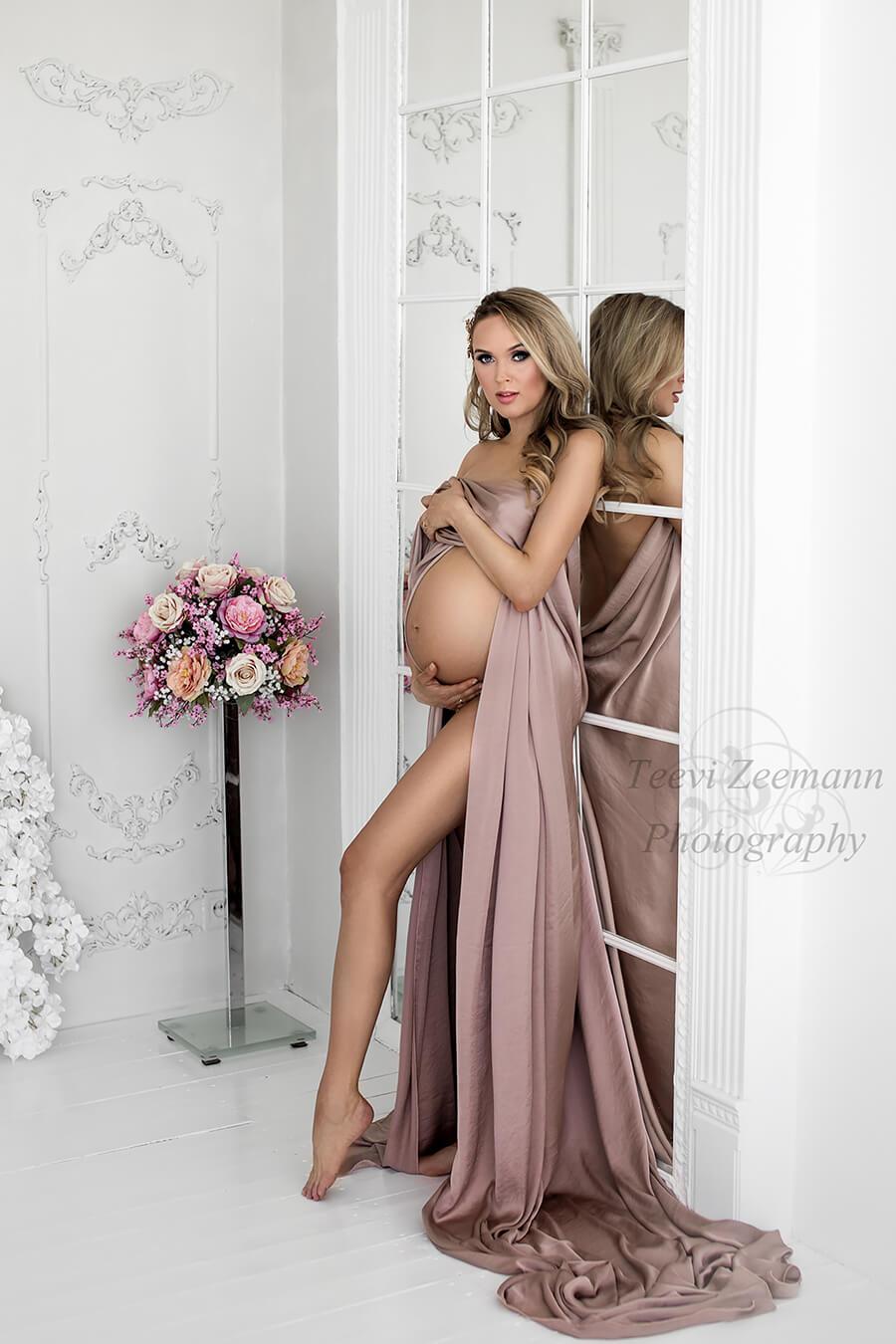 Blond pregnant woman poses against a mirror wall covering her breast with a piece of dusty pink silky fabric. She is staring to the camera and has a flower bouquet next to her.
