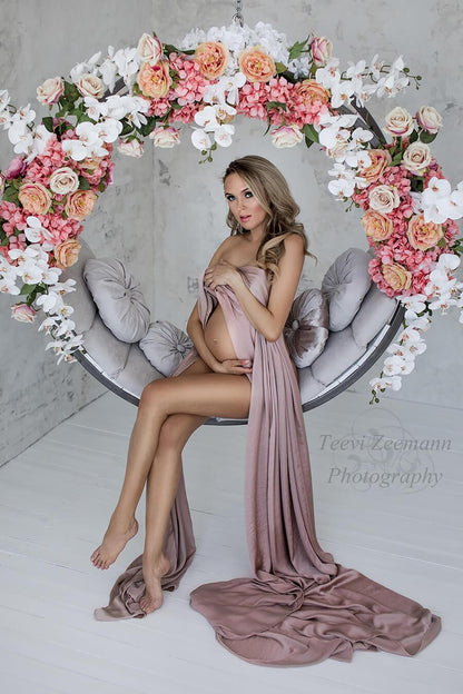 Blond pregnant woman is wearing a silky scarf to cover her breasts. she is sitting on a circle swinger decorated with flowers. She is staring to the camera and her belly and legs are uncovered. 
