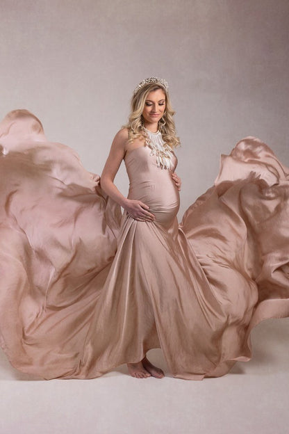 Blond pregnant woman poses wearing a long piece of draping fabric in dusty pink color. The fabric is covering her body and she is also wearing a big feather necklace and a crown. 