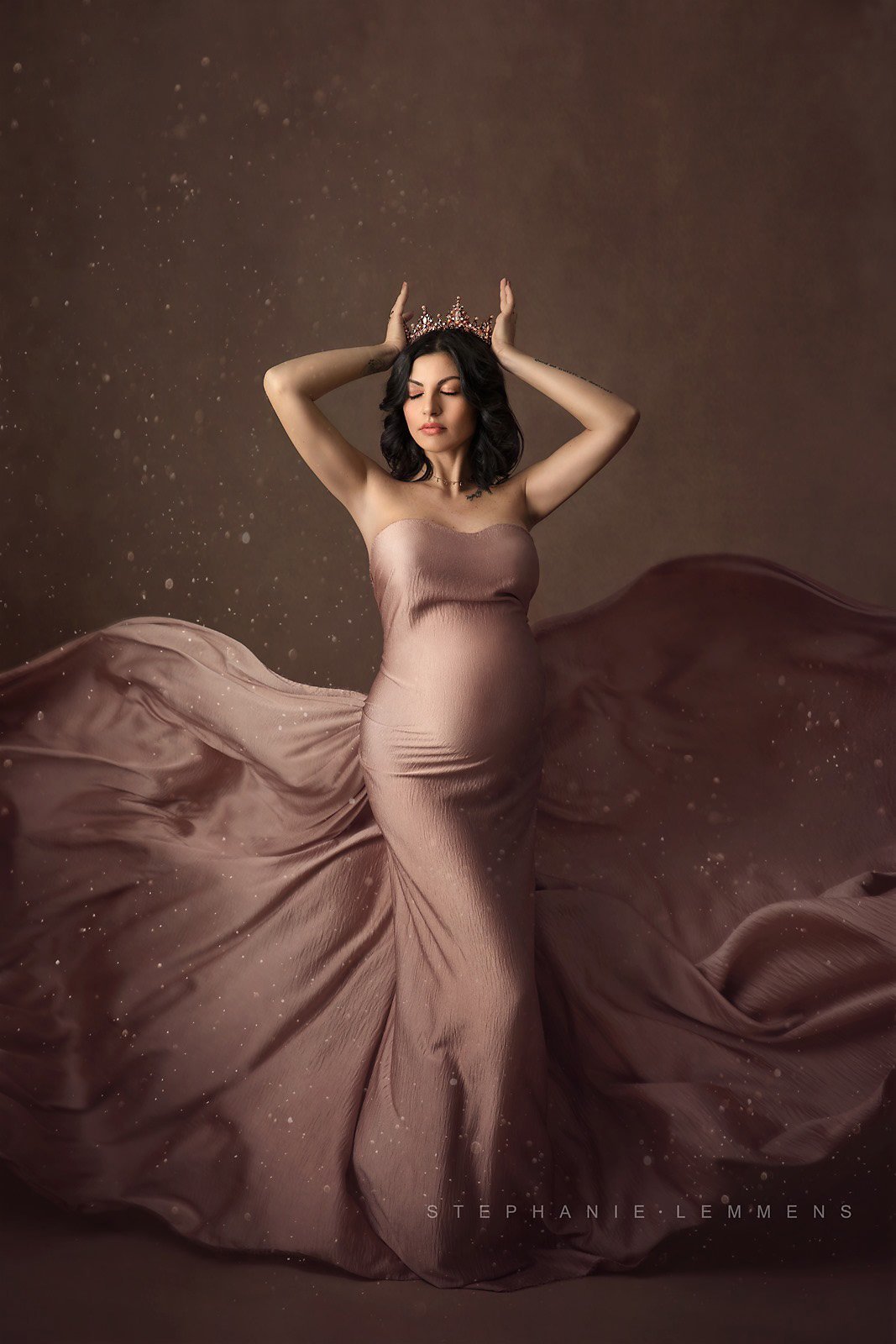 Pregnant model poses in a studio wearing a crown and a draping fabric covering her body. The scarf is dusty pink. Her eyes are closed and she is holding her crown with both hands. 