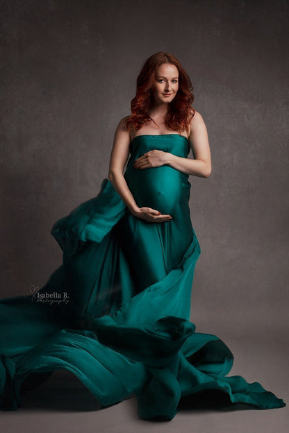 A pregnant woman is wearing a silky scarf as a dress. She is smiling towards the camera. She. has her hands around her belly. The fabric is floating around her at the ground. The color of the scarf is azur.