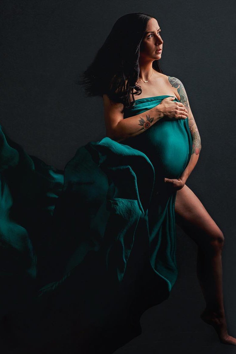 A pregnant woman is posing for a photo. She is holding up the fabric of the silky scarf by her breast. Her other hand is holding the fabric underneath her belly. The rest of the fabric is floating behind her. 