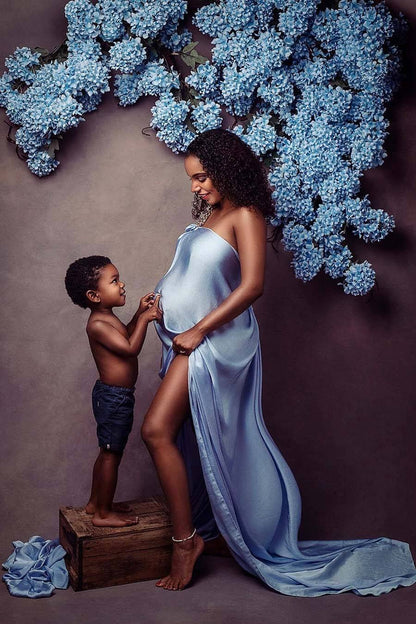 pregnant mother poses together with her son in front of blue hortensias. the mother has a silky scarf in light blue covering her chest and belly. the son is playing with her baby bump and standing on a wooden box. 