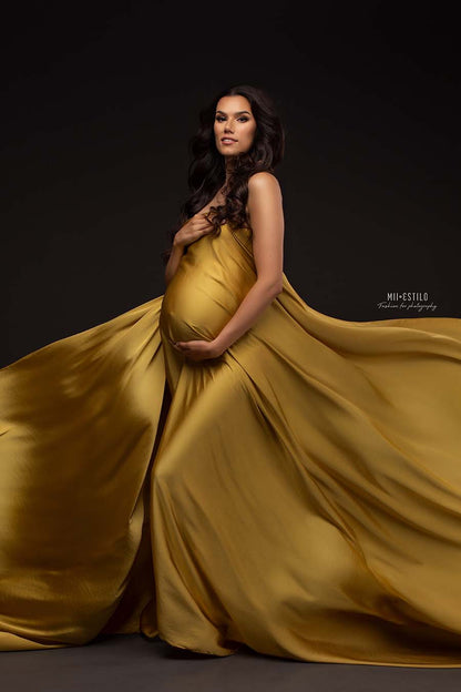 A pregnant model is wearing a silky scar mustard. she is holding the fabric tight around her. The rest of the fabric is being waved next to her. The model has long brown hair.
