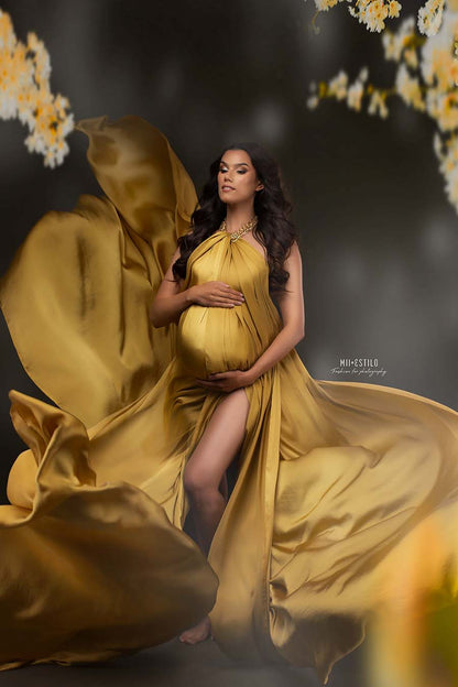 This pregnant model is in the studio. She is wearing the silky scarf mustard as a dress. She has a golden necklace on witch holds up the fabric. They created a split by the leg and flowers around her. The fabric is being tossed around her.