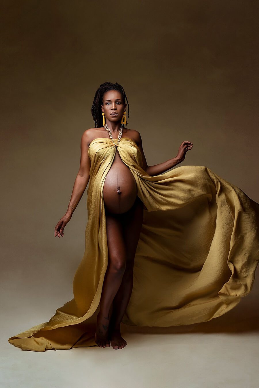 A pregnant model is wearing a silky scarf mustard. The fabric has a shimmer look. The scarf is wrapped like a dress and open at the front. You can see her babybelly. She has her hair in a ponytail. She has tossed the fabric so its stil in the air and gives the photo an extra effect. 
