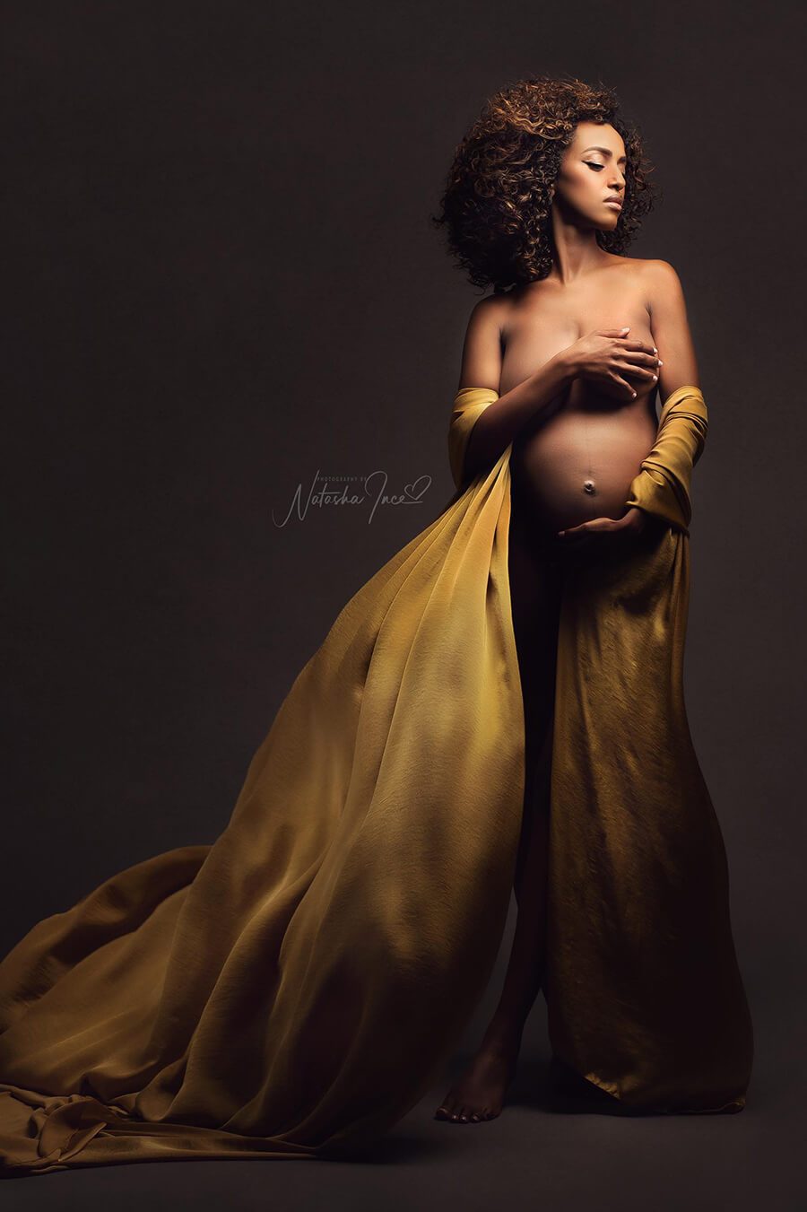 A pregnant model is posing in the studio. She is covering up her breast and has the other hand underneath her belly. The silky scarf mustard is wrapped around her arms and is going down till the ground. 