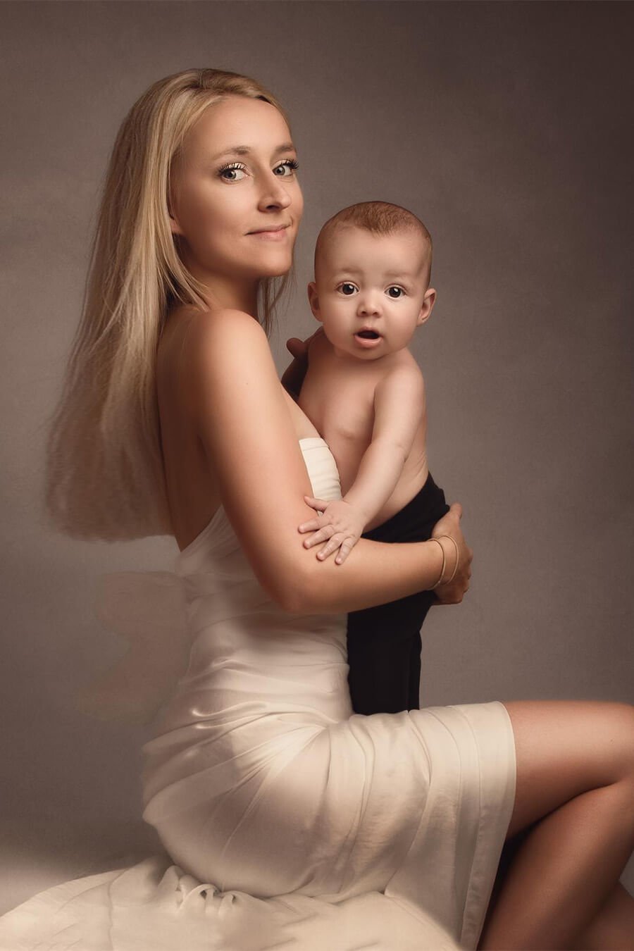 blond model poses with a off white silky scarf wrapped on her body and holding her baby boy on her arms
