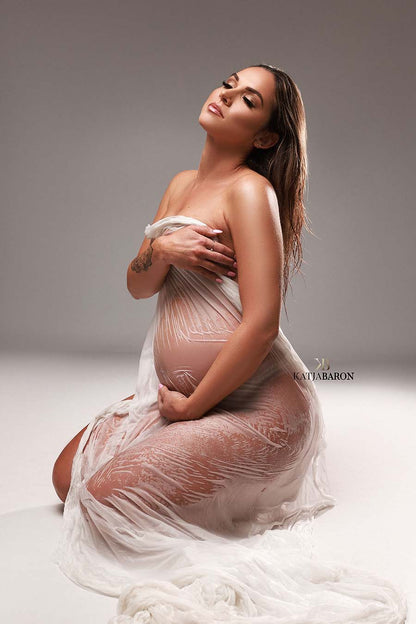 pregnant model poses with a white wet silky scarf covering her body. she is sitting on the studio.