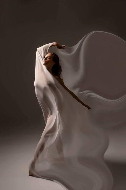 ballerina poses holding a long silky off white scarf against her body. her silhouette can can be seen through the piece. 