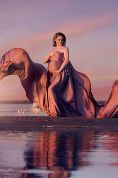 Pregnant model poses by the water wearing a silky draping fabric in old pink. She looks down while covering her body with the fabric. 