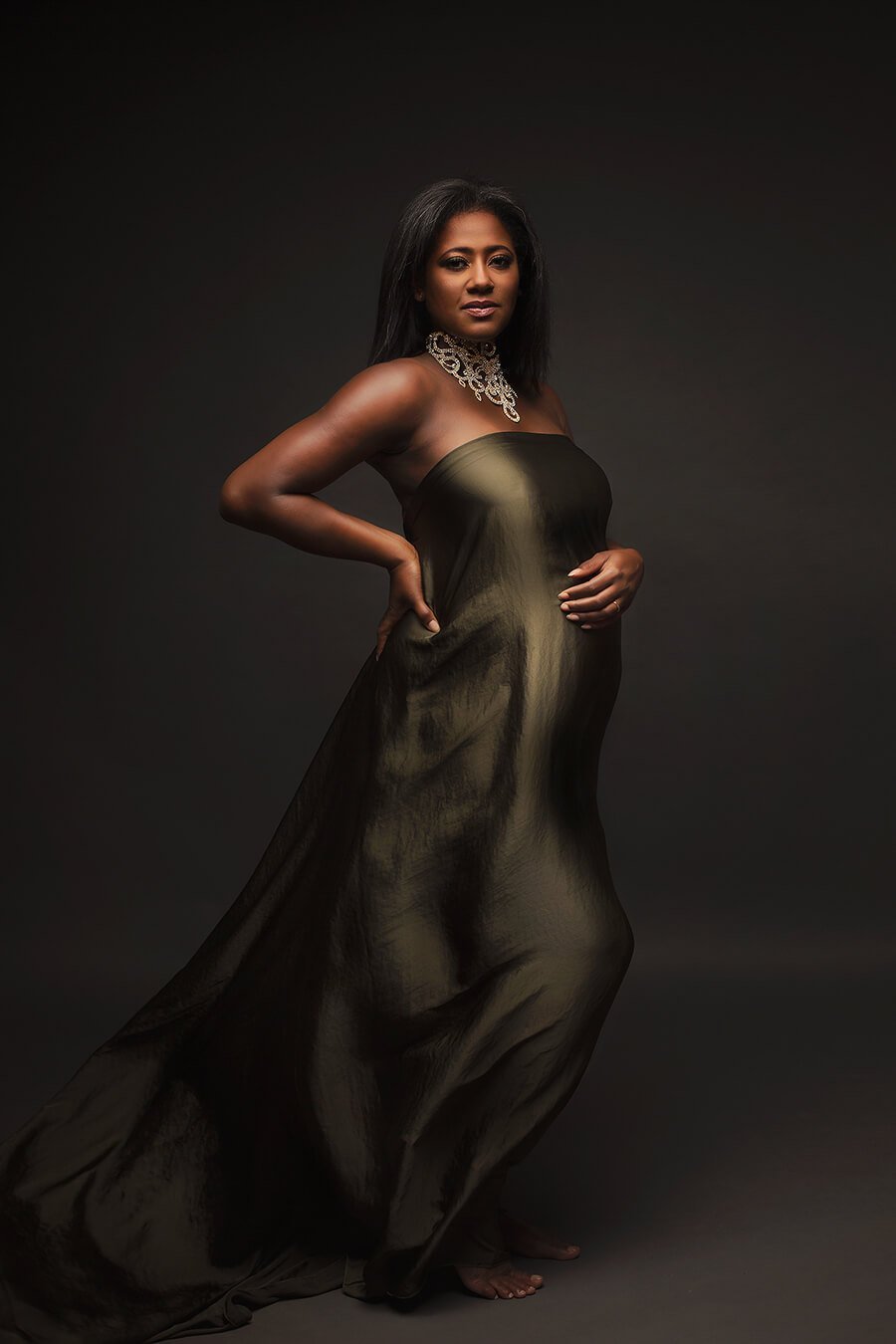 A pregnant model is looking to the camera. She has short black hair. The scarf is around her to cover her up. The scarf is in the color olive and has a silky material for a little bit of shimmer.  The woman is wearing a big white necklace. 